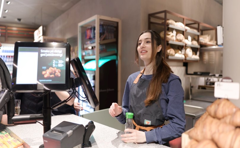 A woman stands at a store counter, engaging with the cashier while surrounded by various products. 