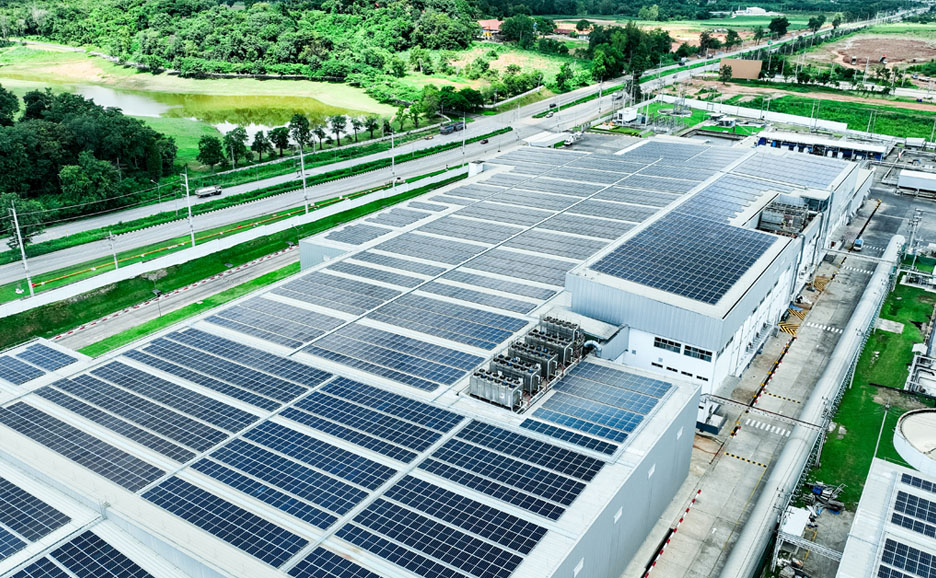 Green factory building with solar arrays installed on the rooftop amidst lush foliage.