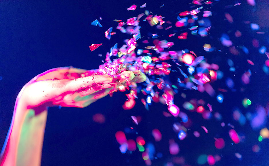 Hand blowing colorful confetti against a dark blue background.
