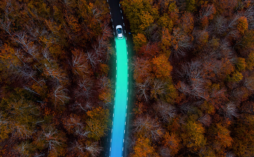 Aerial view of an electric car driving and leaving behind a sustainable trace between autumn forest
