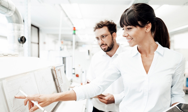 A business scene where two people (a man and a woman) are talking while pointing at a whiteboard
