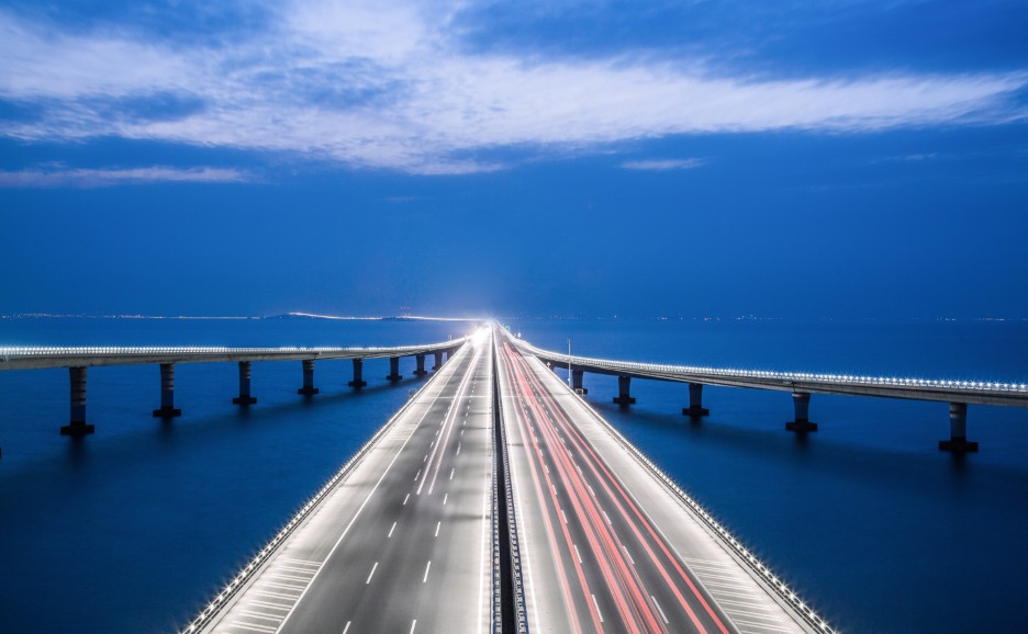 A long bridge stretches across the ocean at night, with streaks of car lights.